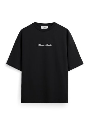 Ung mand med kort hår iført sort oversized T-shirt med teksten "Vortice Berlin" og hvide bukser, stående med hænderne i lommerne mod hvid baggrund.