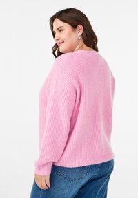 Sieviete ar brūniem matiem, tērpta sudraba auskaros, rozā adītā džemperī un zilās džinsās, nedaudz pagriezusies, rādot džempera aizmuguri.