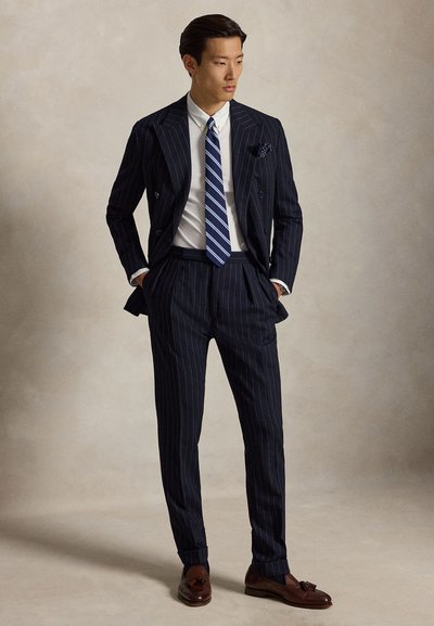 Man draagt een marineblauw krijtstreep pak met wit overhemd, blauw gestreepte stropdas, bruine loafers met kwastjes, staat met handen in de zakken.