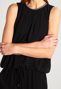 Femme portant un haut sans manches noir avec un col en perles, les bras croisés sur la poitrine devant un fond uni.