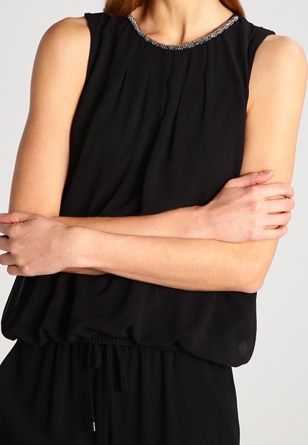 Femme portant un haut sans manches noir avec un col en perles, les bras croisés sur la poitrine devant un fond uni.