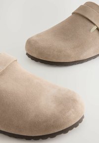 Sabots en suede beige avec un bout arrondi et une semelle en caoutchouc brun plate. Présente une sangle étroite sur le dessus pour un maintien sécurisé.