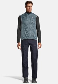 Grauwe leichte Weste über einem langärmeligen schwarzen Oberteil, kombiniert mit marineblauen Cargo-Hosen mit Reißverschlusstaschen und grauen Schuhen.