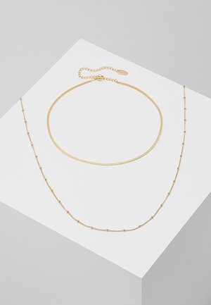 Deux colliers en or délicats sur une surface blanche, l'un avec de petites perles espacées régulièrement et l'autre avec une chaîne lisse et un fermoir réglable.