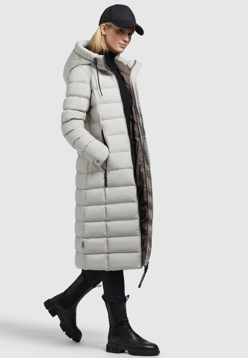 Frau trägt einen langen, hellgrauen, gesteppte Daunenjacke mit Kapuze, schwarzen Rollkragenpullover, Hose, Stiefel und schwarze Baseballmütze vor einem schlichten Hintergrund.