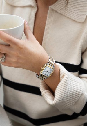Montre rectangulaire en argent et or avec un cadran blanc et des chiffres romains, portée sur un poignet vêtu d'un pull à rayures crème et noir.