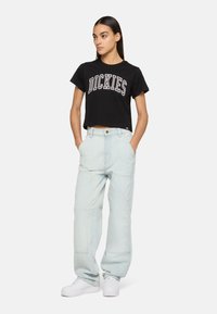 Schwarzes, cropped T-Shirt mit weißem "DICKIES"-Logo, kombiniert mit hellblauen, oversized Hosen und weißen Sneakern. Das Model steht mit den Händen in den Taschen.