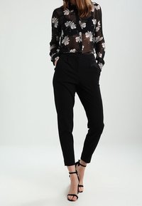 Blusa nera trasparente con stampa floreale, abbinata a pantaloni neri a sigaretta. L'outfit presenta un colletto con bottoni e tacchi con cinturino alla caviglia.
