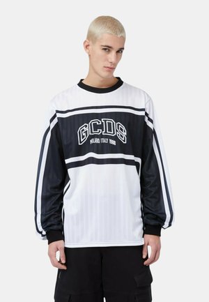 Giovane uomo con capelli biondi corti che indossa una maglia a maniche lunghe bianca e nera con la scritta "GCDS Milano Italia 1988", in piedi davanti a uno sfondo semplice.