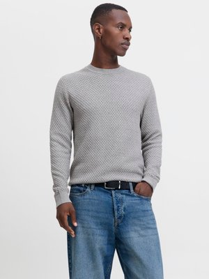 Mann iført en lys grå strukturert genser, blå jeans med svart belte, stående med en hånd i lommen mot en ensfarget bakgrunn.