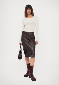Donna che indossa un top bianco a maniche lunghe, una gonna in pelle marrone scuro, stivali bordeaux, mentre tiene una piccola borsa marrone scuro, in piedi davanti a uno sfondo bianco.