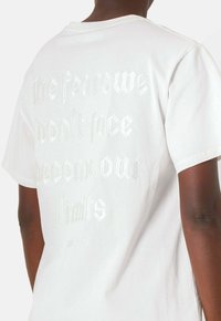 Camiseta de algodón blanca con texto en un estilo de letra plateada en relieve en la parte posterior. Mangas cortas y un ajuste relajado.