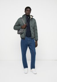Green quilted puffer jacket with a hood, navy sweatshirt underneath, blue denim jeans, and white sneakers. Small gray bag is worn across the body.