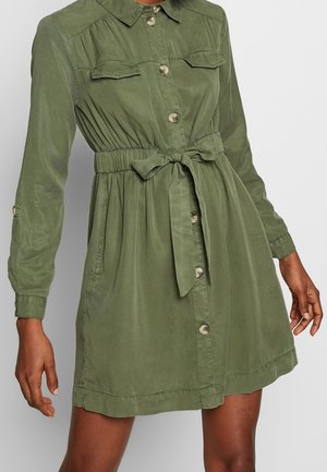 Sukienka koszulowa w oliwkowej zieleni z długimi rękawami, zapięciem na guziki z przodu, dwoma kieszeniami na piersi i paskiem w talii. Gładka tkanina z lekko marszczoną fakturą.