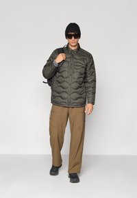 Chaqueta acolchada verde oliva oscuro con cierre de botones, camiseta interior gris, pantalones cargo marrones con bolsillos con cremallera, gorro negro y gafas de sol.
