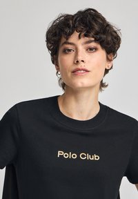 Svart t-shirt med rund halsringning, med "Polo Club" tryckt i guld-bokstäver i mitten. Mjuk, slät tygtextur.