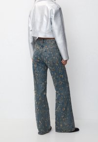 Veste en cuir argentée courte associée à un jean en denim bleu évasé et déchiré, avec des motifs de taches marron clair et des ourlets effilochés.