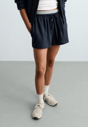 Personne portant un short à fines rayures foncé, des chaussettes blanches, des baskets beiges et une veste sombre avec les mains dans les poches sur un fond uni.