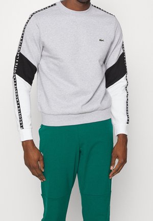 Mężczyzna ubrany w szarą bluzę z czarno-białymi rękawami z logo Lacoste i zielone spodnie sportowe na jednolitym tle.