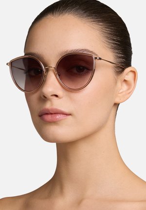 Giovane donna con capelli tirati indietro, che indossa grandi occhiali da sole rosa dorato a forma di occhio di gatto con lenti rosa, espressione neutra e spalle scoperte.