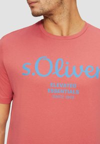 Rosa Baumwoll-T-Shirt mit blauem Drucktext, der "s.Oliver ELEVATED ESSENTIALS SINCE 1969" lautet. Rundhalsausschnitt, kurze Ärmel.