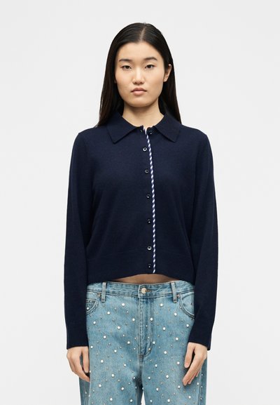 Νεαρή γυναίκα με μπλε σκούρο ζακέτα με κουμπιά πάνω από ριγέ πουκάμισο και ανοιχτόχρωμα τζιν διακοσμημένα με μαργαριτάρια, στέκεται μπροστά σε ένα απλό φόντο.