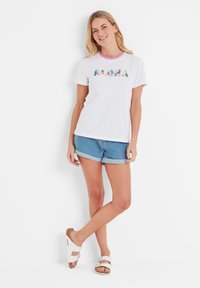White cotton t-shirt with a colorful floral "ALOHA" graphic, short sleeves, and a pink crew neckline, paired with denim shorts and white sandals.