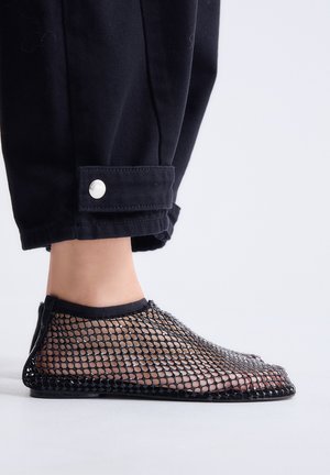 Primer plano de un pie que lleva un zapato negro de malla sin cordones combinado con pantalones negros con un puño con botón a presión por encima del tobillo.