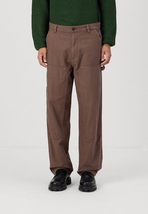 Mann trägt braune gerade geschnittene Hose, schwarze Schuhe und einen dunkelgrünen Strickpullover, steht vor einem einfarbigen hellen Hintergrund.