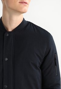 Giubbotto bomber nero con colletto a costine, bottoni a pressione e tasca con zip sulla manica sinistra. Texture del tessuto liscia.