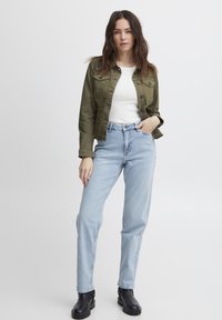 Chaqueta de mezclilla verde oliva sobre una camiseta blanca, combinada con jeans claros de cintura alta y botas negras hasta el tobillo. Diseño simple, estilo casual.