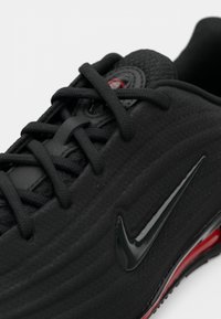 Crni Nike tenisice s crnim vezicama, teksturirana tkanina, sjajni Nike znak i crveno amortiziranje vidljivo na đonu.