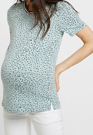 Kobieta w ciąży ubrana w jasnoniebieską, krótko rękawową, wzorzystą bluzkę ciążową i białe spodnie, z jedną ręką podpartą na dole pleców.