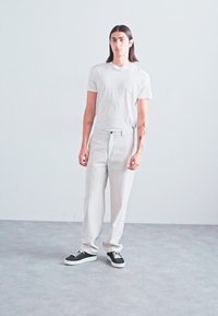 Young person with long dark hair stands wearing white t-shirt, white pants, and black sneakers against plain light gray background.