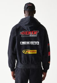 Μαύρο φούτερ με κουκούλα με λογότυπα, ανάμεσά τους "Orbr" και "New Era Racing", σε κόκκινο, λευκό και κίτρινο. Το υλικό φαίνεται μαλακό με ξεθωριαστή υφή.