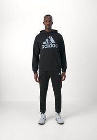 Sudadera negra con gran logo "adidas" en azul claro, combinada con pantalones de chándal negros. Calzado deportivo gris claro con acentos oscuros, textura mínima.
