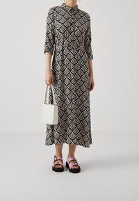 Maxime dressi mustalla ja valkoisella lehtikuviolla, pitkät hihat ja kaulus. Yhdistettynä pieneen valkoiseen käsilaukkuun ja vaaleanpunaisiin sandaaleihin.