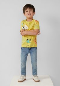 Gelbes Baumwoll-T-Shirt mit verschiedenen gedruckten Emojis und Designs; kombiniert mit blauen Denim-Jeans und weißen Sneakers mit beigen Akzenten.