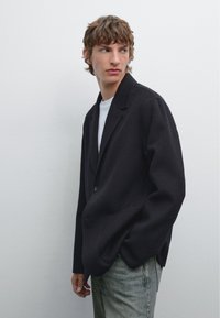 Black wool blazer with a single button, notched lapels, and side pockets, paired with a white t-shirt and light denim jeans.