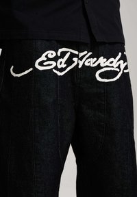 Close-up of black denim pants with white "Ed Hardy" cursive text across the upper thigh area.