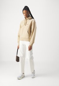 Beige hooded sweatshirt with a front pocket and embroidered logo, paired with white pants and grey sneakers. Small animal-print bag included.