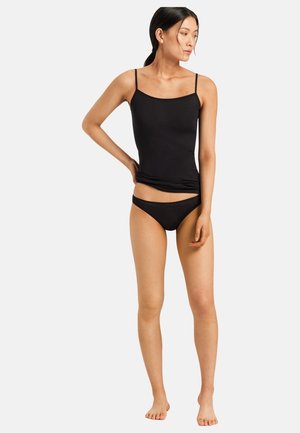 Pitkätukkainen nainen, jolla musta camisole ja mustat alusvaatteet, seisoo paljain jaloin yksinkertaisen vaalean taustan edessä.