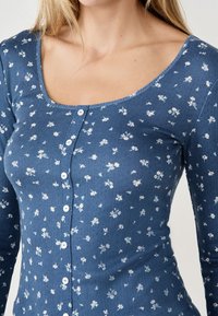 Niebieska bluzka na długi rękaw w kwiatowy wzór, wykonana z prążkowanej tkaniny, z dekoltem w serek i białymi zapięciami na guziki z przodu.