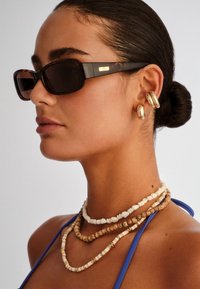 Γυναίκα με σκούρα μαλλιά συλλεγμένα σε κότσο φορά γυαλιά ηλίου με σχέδιο χελώνας, χρυσούς κρίκους σκουλαρίκια και πολυεπίπεδα κολιέ από χάντρες με μπλε τοπ με τιράντες.