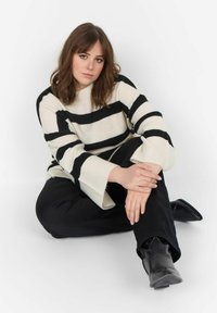 Cremfarbener und schwarzer gestreifter Strickpullover mit weiten Ärmeln, kombiniert mit schwarzen Hosen und spitzen schwarzen Stiefeln. Modell sitzt vor einem weißen Hintergrund.