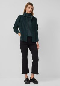 Femme debout portant une chemise boutonnée vert foncé par-dessus un col roulé assorti, un pantalon cropped noir et des mocassins en cuir verni noirs.