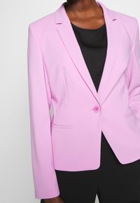 Svetle růžový blejzr vyrobený z hladké tkaniny, se zářezovými klopami, jedním knoflíkem a dvěma předními kapsami. Černý top pod ním.