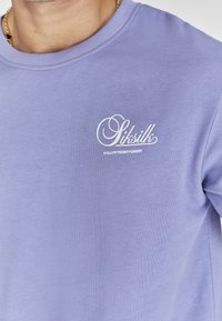 Gai purpursarkans džemperis no mīksta materiāla, ar baltu logotipu un tekstu uz krūtīm. Ribuveida apkakle un īsās piedurknes.