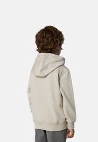 Sudadera beige con una gran capucha, puños y dobladillo acanalados. Material de algodón suave. Niño de espaldas, mostrando el diseño y la textura de la parte trasera.