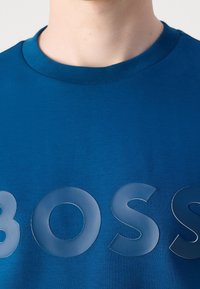 T-shirt bleu col rond avec de grandes lettres brillantes épelant "BOSS" sur la poitrine, porté par une personne avec un cou et une mâchoire visibles.